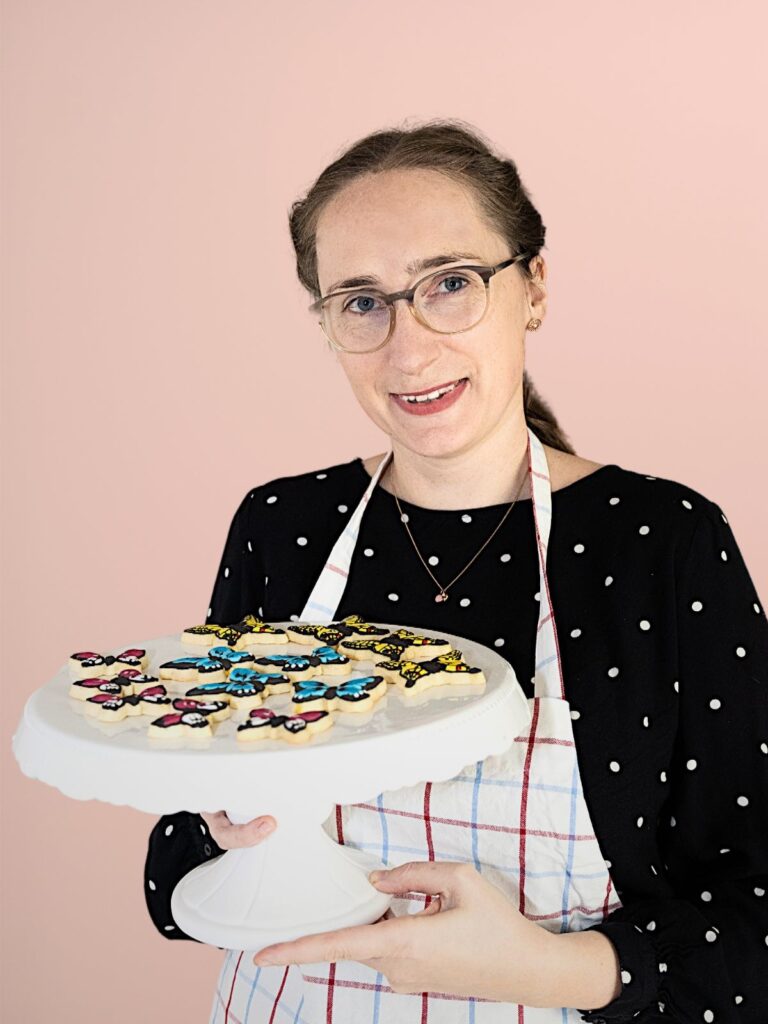Eine lächelnde Bäckerin, blickt mit einer Auswahl ihrer liebevoll dekorierten Kekse in die Kamera.